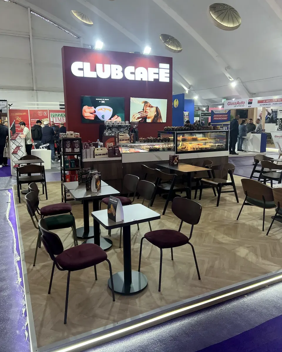 Vue d’ensemble du stand Club Café : mur de marque, comptoir, tables et chaises sur un sol décor parquet